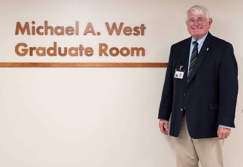 Michael West poses in front of the sign of the Michael A. West Graduate Room on the fourth floor of Memorial Library.